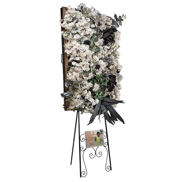 تابلوگل تسلیت ماه ملک از موسسه خیریه مهربانان، شامل استند فلزی مشکی با طرحهای اسلیمی، کانوپی چوبی در بالا و پایهای برای قرار دادن عکس یا متن در پایین. تابلوگل با گلهای انبوه سفید، چند برگ تیره و گلهای مخروطی مشکی تزئین شده است. عواید حاصل از اجاره این تابلوگل صرف امور خیریه میشود.