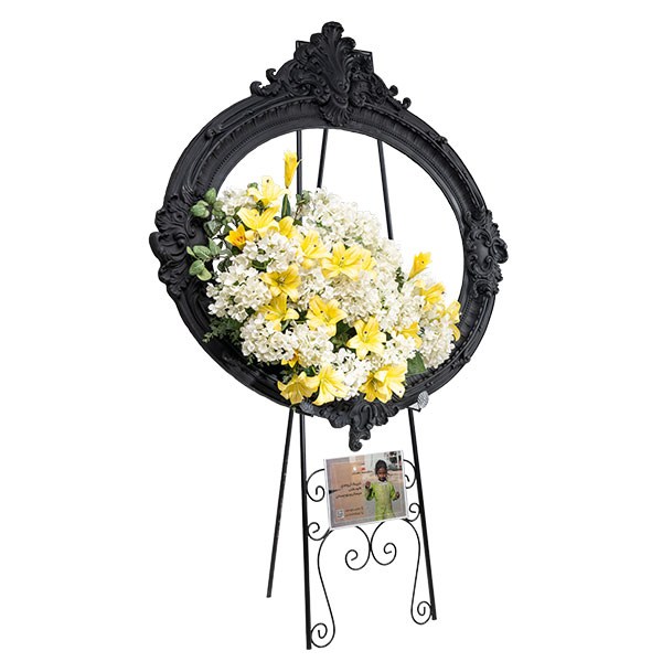 تابلوگل تسلیت مدل هانل با قاب بیضی سفید منبتکاری شده و گلهای رز صورتی و شکوفههای سفید، قرار گرفته بر روی یک سهپایه فلزی طلایی رنگ